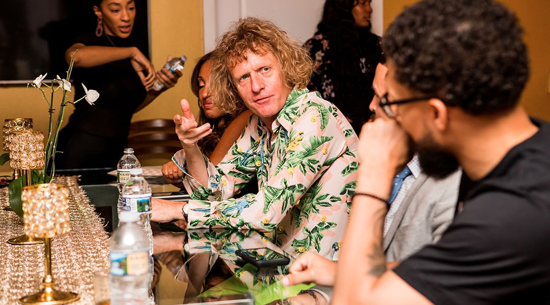 Grayson Perry sitting at a bar wearing a patterned shirt having a conversation with the individual on his left